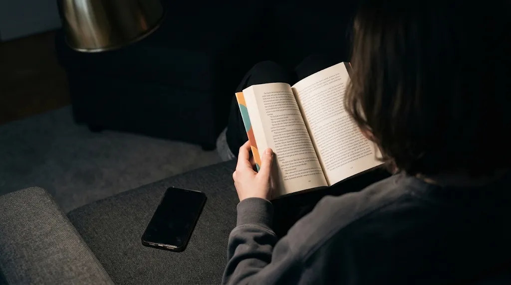 Over the shoulder of someone on a sofa reading a paperback, their phone set aside next to them